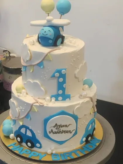 A charming two-tier cake for a first birthday celebration. The design includes a cute helicopter topper, cars, and fluffy clouds against a clean white background.