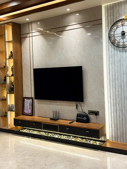 A close-up of a sophisticated TV entertainment unit. The design includes a pebble-filled base with ambient lighting, a sleek console, and a backlit display shelf, all set against a marble and fluted panel wall.