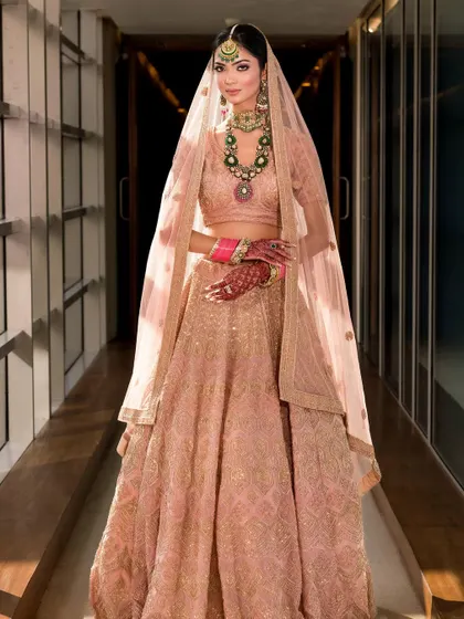 A breathtaking full-length shot of a soft peach-pink lehenga, captured in a way that feels both grand and intimate. The light beautifully catches the sheer dupatta.
