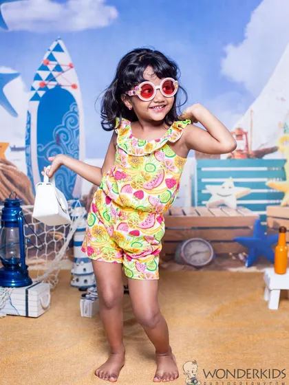 A little girl strikes a pose in her colorful summer outfit against a fun beach backdrop. Her confident smile and stylish sunglasses make this a super fun and vibrant kid's portrait.