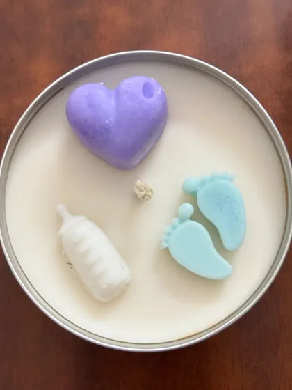 This baby shower candle in a tin container is topped with cute, baby-themed wax elements like a heart, a milk bottle, and tiny feet. All the toppings are made of soy wax.