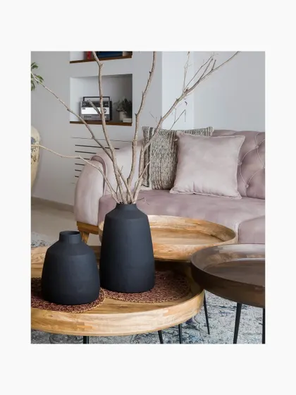 A close-up of the nested wooden coffee tables, topped with matte black vases and dried branches. The soft pink sofa in the background adds a gentle warmth to the composition.