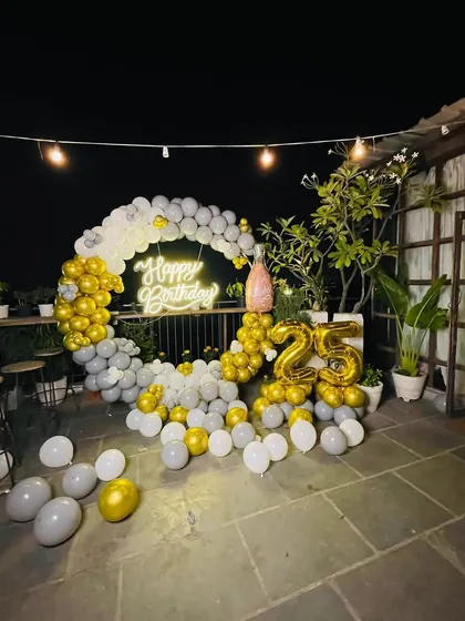 An elegant 25th birthday party on a terrace. The decor features a white and gold circular balloon arch, a neon sign, and matching foil balloons for the age.