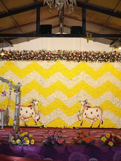 This full-stage view shows a beautiful floral wall with a yellow and white chevron pattern. The design includes traditional cow motifs and a decorated cradle, making it suitable for a combined Seemantham and naming ceremony.