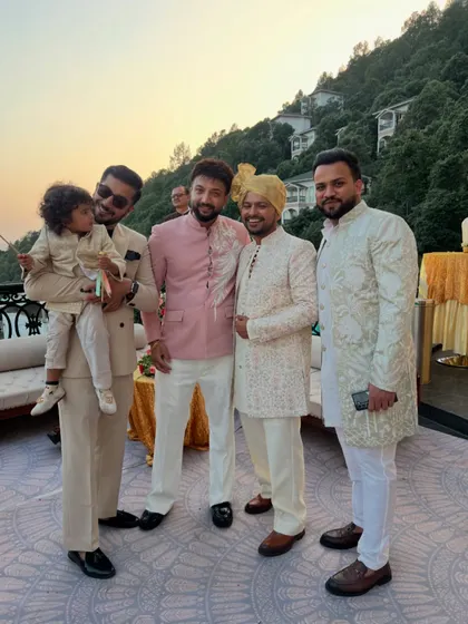 A group of friends and family at a wedding, dressed in a variety of our custom outfits. The collection includes a matching father-son suit, a pink Jodhpuri jacket, and embroidered sherwanis.
