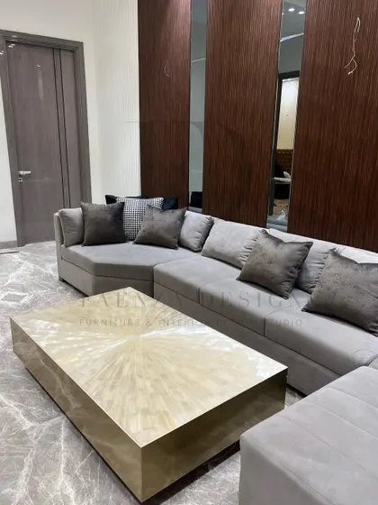 A spacious living room designed for comfort and style. The large grey sectional sofa is complemented by a statement coffee table with a brass finish, set against a backdrop of wood and mirror wall paneling.