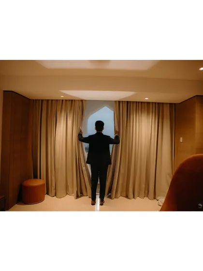 An artistic shot of the groom looking out the window, framed by curtains. This creates a sense of anticipation and quiet reflection.