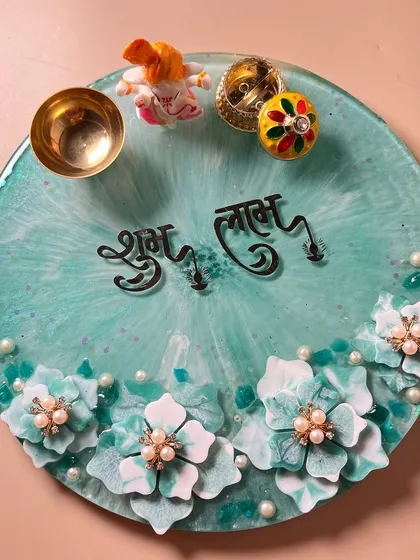 Bring a touch of handmade beauty to your prayers. This is a custom 10-inch resin pooja thali in a lovely sea-green shade, decorated with handcrafted flowers and pearls. It comes with a matching diya, Ganesh idol, and a small bowl for offerings.