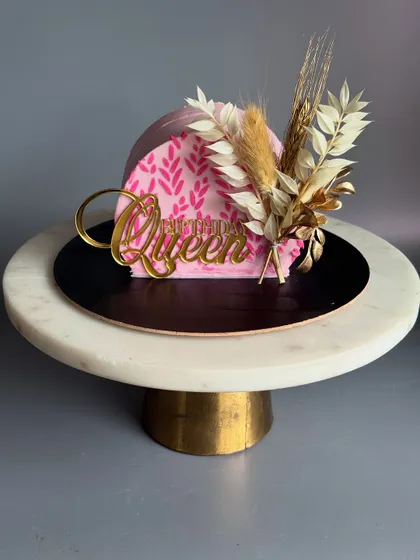 A chic and modern cake-top-forward design. This small pink cake is adorned with a stencil pattern, dried flowers, and a gold 'Birthday Queen' topper, perfect for an intimate celebration.