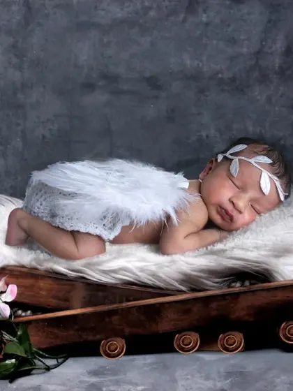 A baby boy with angel wings sleeping on a rustic prop against a textured grey background.
