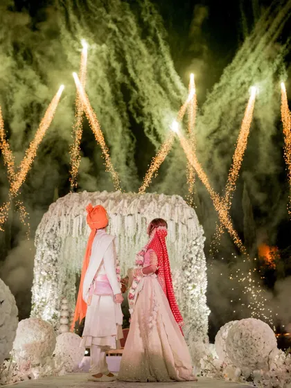 A moment frozen in time, where love and light collide. The couple stands before a floral mandap as fireworks paint the sky, a signature of my grand wedding planning.