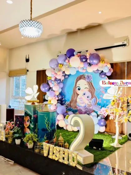 This wide shot shows a large-scale mermaid party setup on a stage, complete with marquee numbers, custom printed pedestals, and themed props.