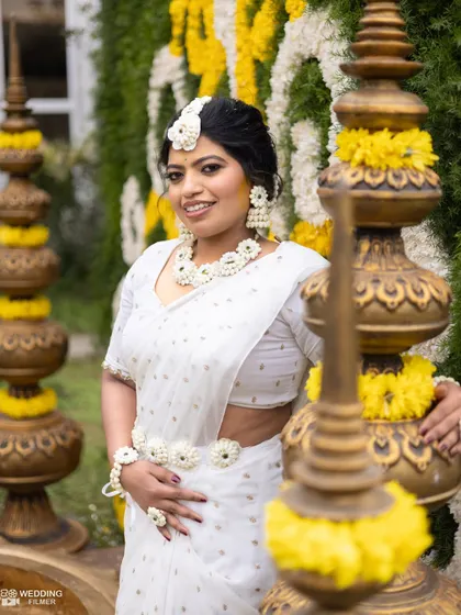 The bride, dressed in an elegant white saree with floral jewelry, poses gracefully amidst traditional Haldi decor.