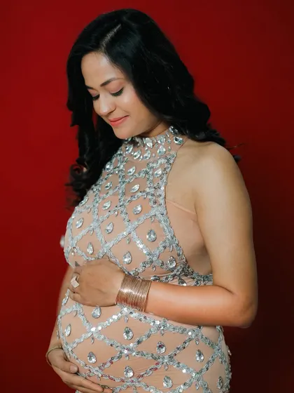 A close-up portrait highlighting the 'pregnancy glow'. The mother-to-be looks down at her bump, wearing a stunning, jewel-encrusted gown against a rich red backdrop.