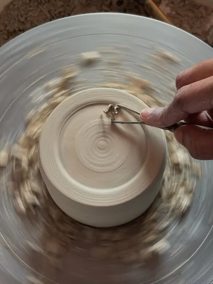 Trimming the base of a leather hard pot. This is a critical step to remove excess clay and create a finished foot ring, giving the piece stability and a professional look. The spinning wheel creates a beautiful spiral of clay shavings.