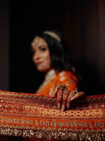 An artistic shot focusing on the bride's hand, adorned with henna, resting on her ornate dupatta. The inscription 'Sada Saubhagyavati Bhava' is visible, adding a traditional blessing.