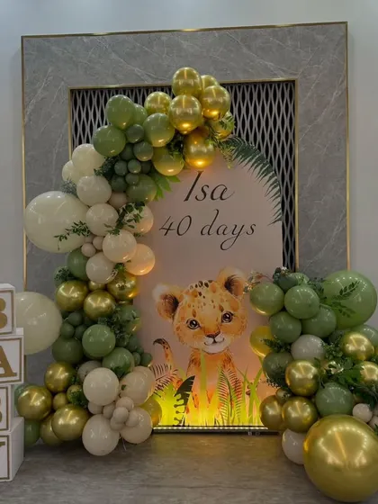 A cute baby lion theme for a '40 days' celebration. This setup includes a personalized backdrop, a balloon garland in green and gold, and 'BABY' display boxes.