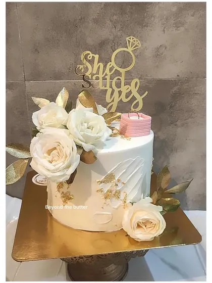 A classic "She Said Yes" engagement cake. The clean white design is elevated with gold-painted leaves, fresh white roses, and a miniature ring box, making it perfect for celebrating the proposal.