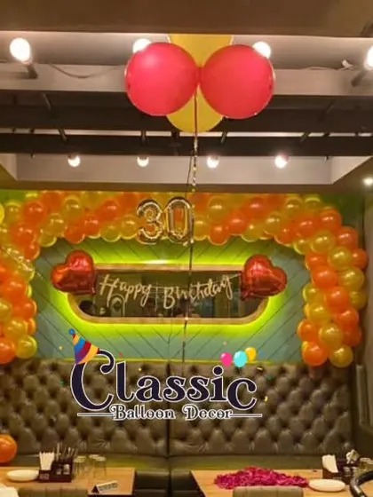 A full restaurant booth decorated for a 30th birthday, with an orange and yellow balloon arch and a neon-lit backdrop.