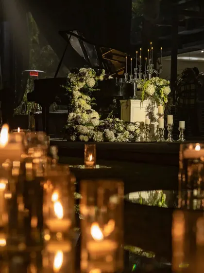The glow of countless candles reflects off the polished surface of the grand piano, which is adorned with a lavish floral display. This scene captures the romantic and dramatic essence of the black and gold cocktail night.