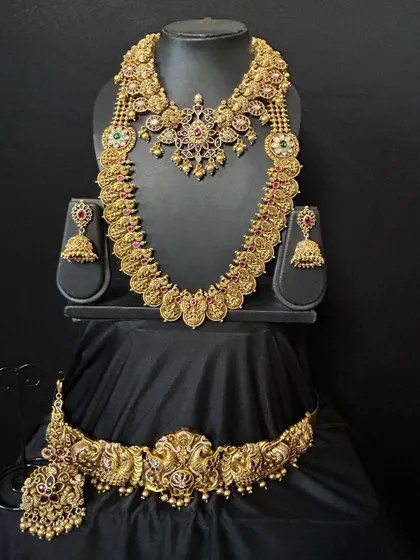 This stunning temple jewellery set combines a short necklace, a long coin haaram, and an intricate vaddanam. The craftsmanship is evident in the detailed deity motifs, making it a truly divine choice for your wedding day.