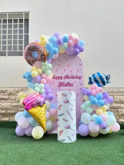 A pastel candy-themed birthday party for an outdoor setting. The decor includes a custom backdrop and foil balloons shaped like ice cream, donuts, and candy.