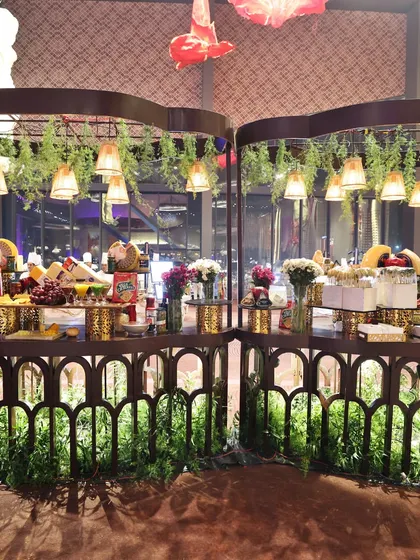 A rustic-themed cheese and charcuterie station, decorated with hanging greenery and pendant lights. The presentation is both charming and abundant, creating a warm and inviting atmosphere.