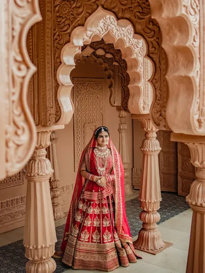 A stunning bridal portrait framed by ornate Indian archways. The bride, in her traditional red lehenga, looks like royalty in this grand architectural setting.