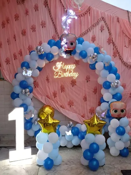 A fun Boss Baby themed first birthday party. The setup includes a balloon arch in blue and white, a "Happy Birthday" neon sign, a light-up number "1," and Boss Baby character balloons.