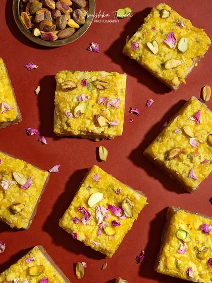 A flat lay of Sutarfeni, a traditional Diwali sweet, with pistachios and rose petals scattered on a rich red background.