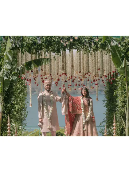 The couple stands united under a magnificent mandap of cascading white flowers and lush greenery, with a serene lake visible in the background.