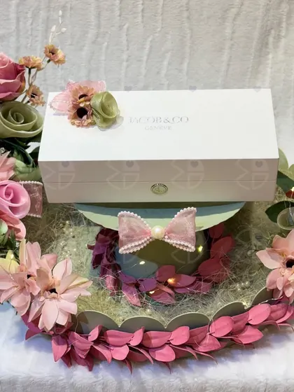 A beautiful gift platter for an engagement or Roka. A luxury watch box is presented on a tray adorned with a delicate arrangement of pink and green artificial flowers.