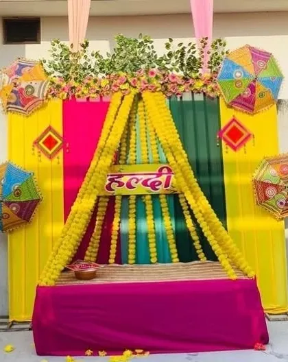 A canopy-style seating for a Haldi ceremony, created with strings of marigold flowers over a pink diwan. The backdrop is a mix of bright yellow, pink, and green drapes.