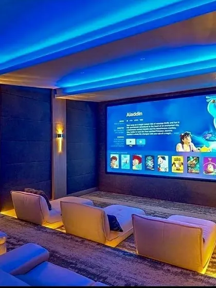 This contemporary media room is designed for both family enjoyment and critical viewing, featuring modern lounge seating with integrated floor lighting. The large-format screen is complemented by blue ambient cove lighting, creating an inviting atmosphere while ensuring excellent picture quality for everything from animated films to blockbuster movies.