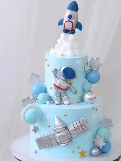 A two-tiered astronaut and rocket ship cake for a space adventure. This light blue cake is decorated with planets, stars, and a satellite, perfect for a future space explorer.
