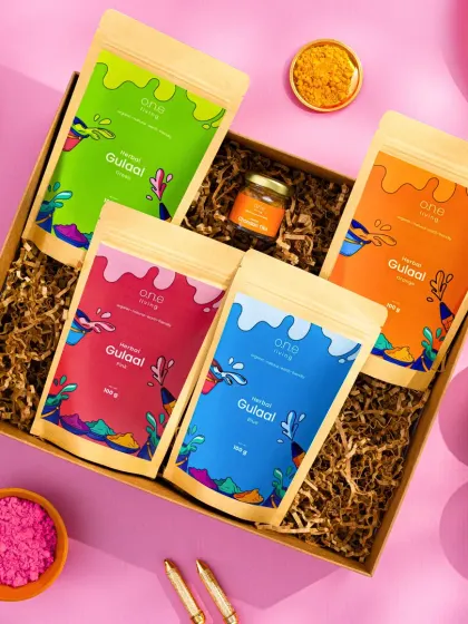 A top-down shot of One Living's herbal gulal box. The bright pink background and scattered colors create a playful and festive mood, perfect for the Holi season.