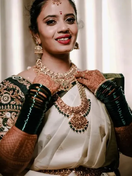 A smiling bride in a traditional white and green saree, adorned with beautiful gold jewelry and dark green bangles.