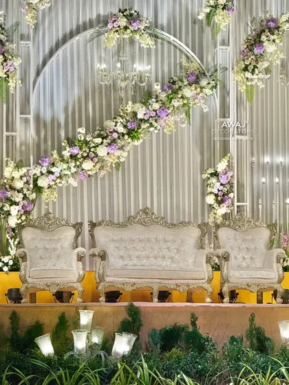 A modern and sophisticated stage design featuring a sweeping arc of white and lavender florals over ornate silver thrones, set against a contemporary silver-paneled backdrop.