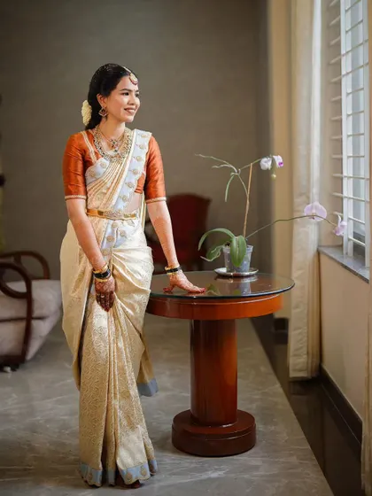 A beautiful, naturally lit portrait of a bride in a classic off-white and rust-orange silk saree. The makeup is kept soft and natural to enhance her features, creating a look of pure elegance.