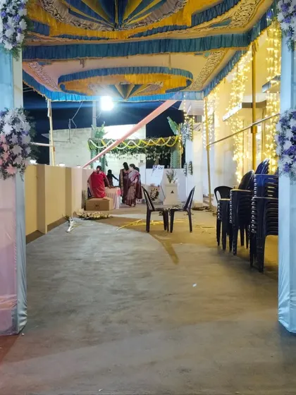 For larger housewarming celebrations, I can arrange for outdoor pandals. This setup features a colorful fabric ceiling in blue and yellow, with pillars wrapped in white cloth and decorated with elegant floral bouquets to accommodate more guests comfortably.