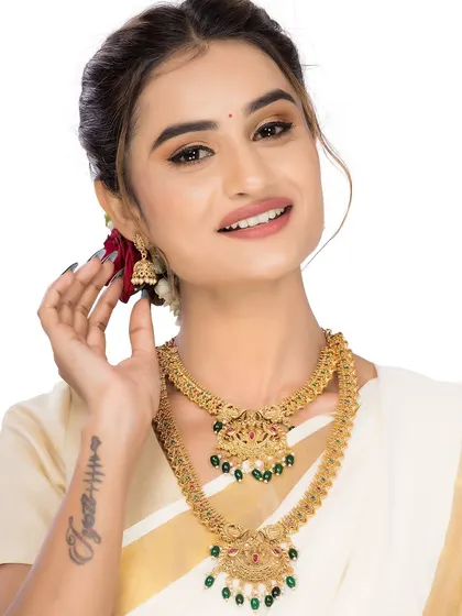 A cheerful model displaying a South Indian temple jewellery set, featuring intricate gold work and green stones, against a clean white background.