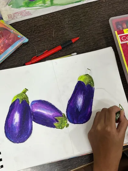 A student uses oil pastels to draw brinjals, learning blending techniques to show highlights and shadows. We teach various mediums as required by the elementary and intermediate exam curriculum.