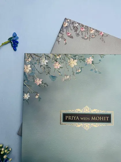A close-up of a pastel blue invitation adorned with delicate, embossed white and pink flowers, creating a soft and romantic feel.