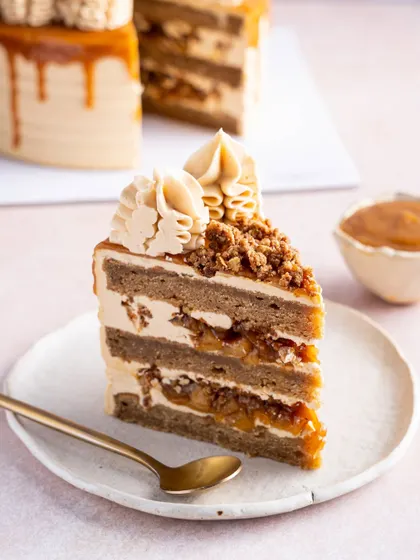 A close-up of a slice of our Apple & Salted Caramel Cake, highlighting the distinct, generous layers. The spiced apple filling and crumble are clearly visible, promising a dessert full of texture and warmth.