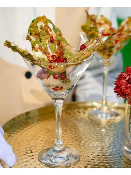 A creative vegetarian appetizer of Palak Patta Chaat served elegantly in a martini glass, combining crispy spinach leaves with tangy chutneys and pomegranate seeds.