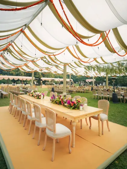 An elegant setup for a wedding lunch. Long wooden tables are set under a billowy canopy with olive and white drapes, accented with marigold garlands for a touch of tradition.