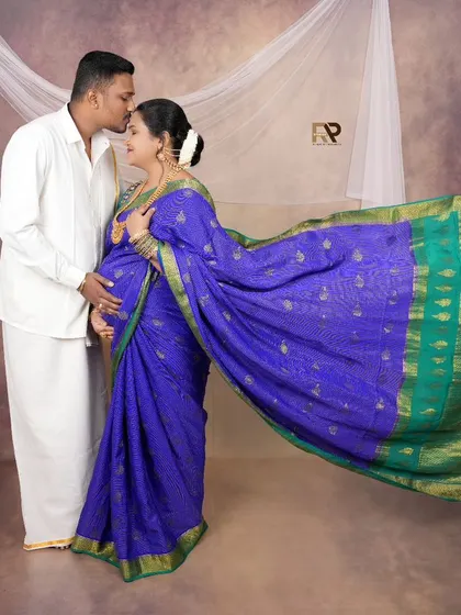 A tender moment in traditional attire. The dad-to-be, dressed in a classic white dhoti and shirt, gently kisses his partner's forehead. The flowing pallu of her blue saree adds a touch of drama.