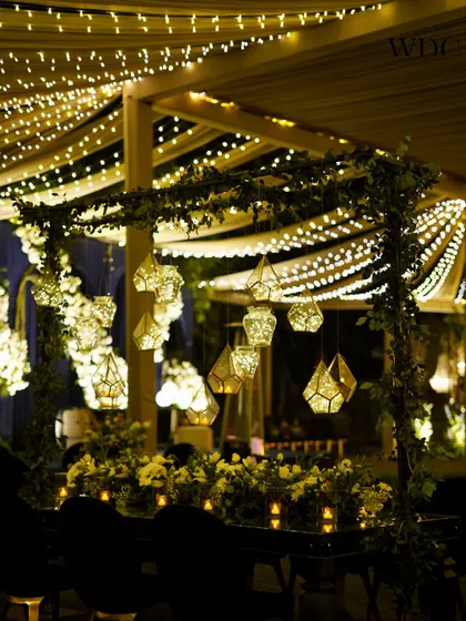 An intimate dining setup under a canopy of fairy lights. The table is adorned with geometric hanging lanterns and a runner of white flowers, perfect for a cocktail party.