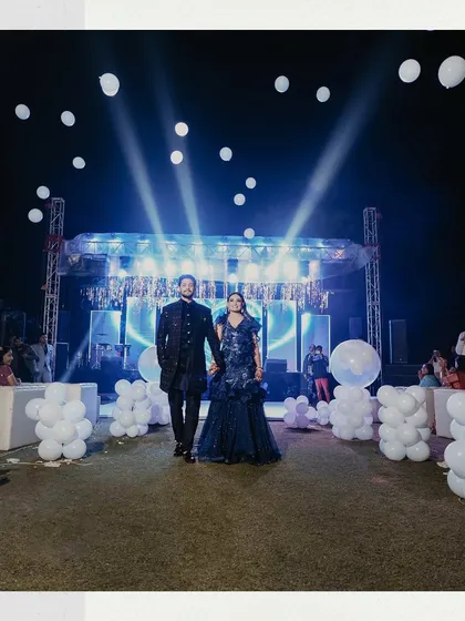 A wide shot of the couple making their entrance at the Sangeet. The large stage, lighting, and balloon decorations highlight the grand scale of the celebration.