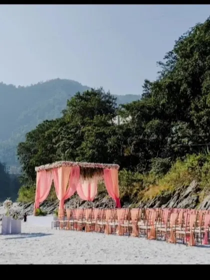 A wide shot of a destination wedding setup on a riverbank. The pink-draped mandap and guest seating look beautiful against the backdrop of the mountains and river.